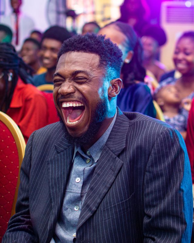 Portrait of a smiling man in a suit laughing amidst a vibrant indoor gathering in Makurdi, Nigeria.